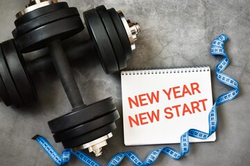 Dumbbells, measuring tape, notepad with text message NEW YEAR NEW START on gray background.