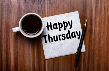 On a wooden table next to a white cup of coffee and a pen is a white paper napkin with the words HAPPY THURSDAY
