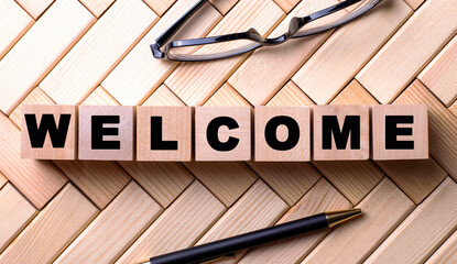 The word WELCOME is written on wooden cubes on a wooden background next to a pen and glasses.