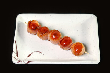 shish kebab from Cherie's tomatoes wrapped in bacon on a plate it is isolated on a dark background