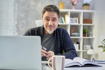 Bearded man working online with laptop computer at home sitting at desk. Home office, browsing internet, study room. Portrait of mature age, middle age, mid adult man in 50s.