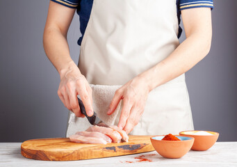 Close up image of a woman chef slicing chicken breast to male stripes and nuggets. She cuts the meat on wooden chopping board using sharp knife. Cayenne red pepper and salt are on side for seasoning