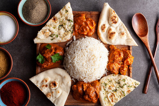 Flat lay image of traditional Indian cuisine platter on dark background with spices and herbs on the side. Assortment contains naan breads as well as rice, butter chicken, paneer, curry, tikka masala