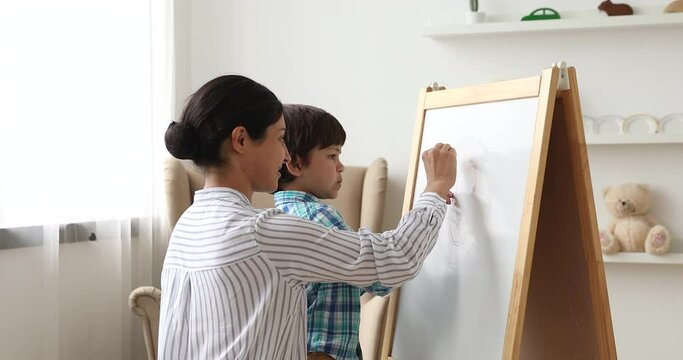 Affectionate young indian ethnicity mother helping or teaching small preschool cute child son drawing on white board with colored crayons. Happy family enjoying creative weekend pastime at home.