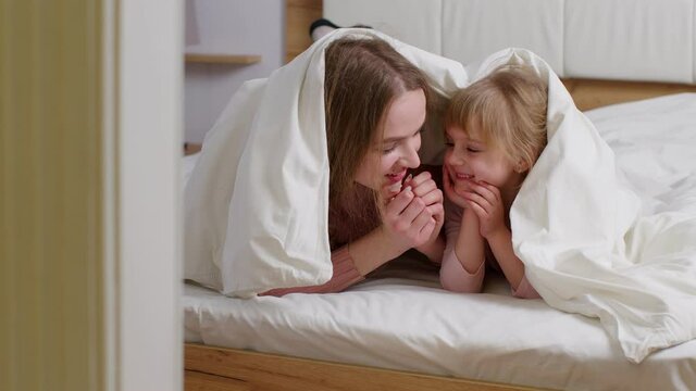 Happy family of mother with daughter lying on bed under duvet blanket, telling secrets to each other