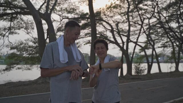 senior asian couple sharing good moment together when outdoor workout at park in evening sunset. happy elderly lover looking at sport smart watch information and proud. smile mature adult exercising.