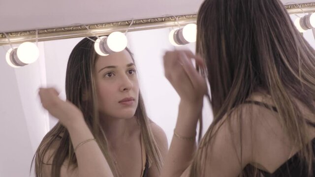 Model examining her face by looking into a lighted makeup mirror in the studio. Actress preparing for a show. Woman with long straight hair standing in front of a mirror and looking at herself.