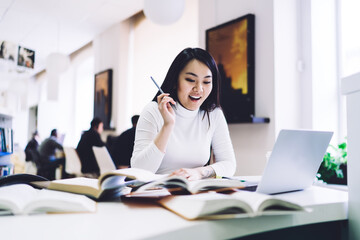 Elegant adult ethnic woman rejoicing breakthrough while sitting at laptop with notepad and books in office