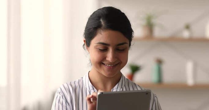 Close up head shot smiling young indian ethnicity woman using touchscreen digital computer tablet, using software apps, web surfing reading interesting information or communicating distantly online.