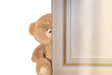 A cheerful toy brown bear peeks out from behind a wooden door.Copy space, space for text on a white and wooden background