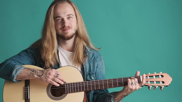 Attractive long haired blond man dressed in casual wear playing on acoustic guitar over blue background. Musician with guitar in studio. Music concept