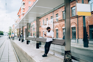 Stylish male hipster leaning on tram station while messaging on smartphone