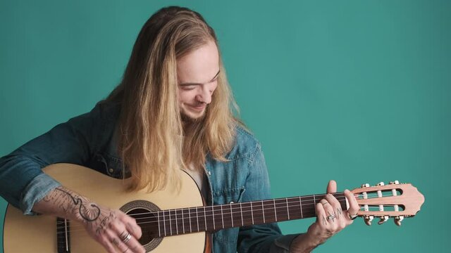 Attractive long haired blond guy looking happy playing on acoustic guitar preparing to perform isolated on blue background. Music concept