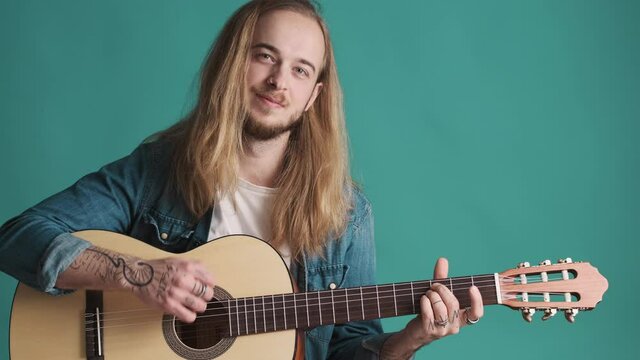 Attractive long haired blond guy playing on acoustic guitar looking concfident on camera over colorful background. Musician with guitar in studio