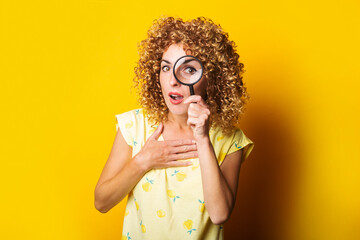 surprised curly girl looks through a magnifying glass on a yellow background.
