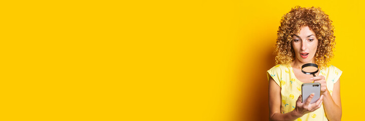 shocked curly young woman looking with a magnifying glass into the phone on a yellow background....