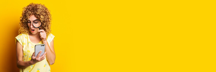 shocked curly young woman looking with a magnifying glass into the phone on a yellow background....