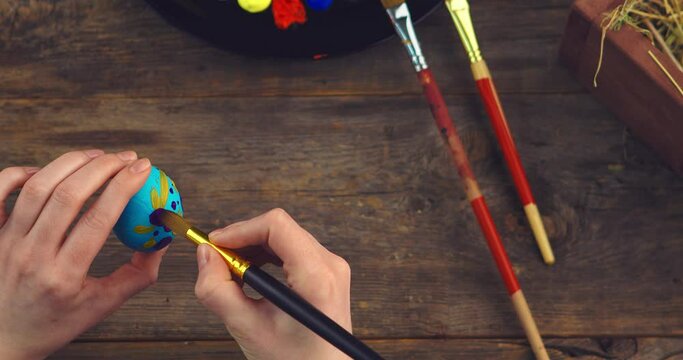 Artist's female hands painting patterns on Easter egg on wooden surface. Top view close up video for christian holiday