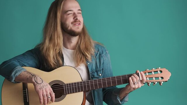 Young long haired blond musician looking inspiring playing on acoustic guitar and singing new song over blue background. Music concept
