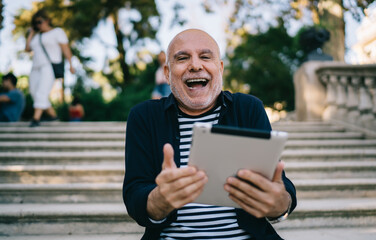 Half length portrait of cheerful senior traveller with modern touch pad gadget laughing at camera during free time in city, excited male tourist 60s with digital tablet rejoicing on retirement