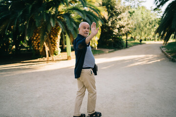 Full length portrait of typical Italian citizen waving during walk daytime in public park with palm trees, casual dressed male senior in comfort outfit clothing looking at camera while gesturing