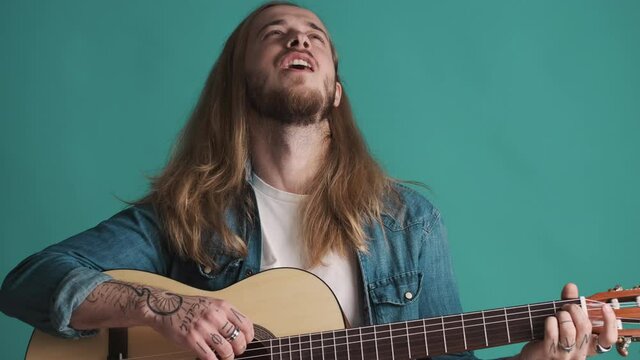 Cool long haired blond bearded musician playing on guitar and singing song loudly over colorful background. Music concept