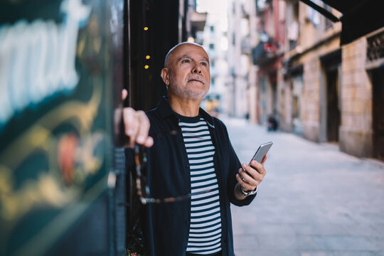 Contemplative Male Thoughtful Looking Away While Waiting For Roaming Notification On Smartphone, Pensive Aged Man With Cellphone Technology In Hand Thinking About Network Blogging On Retirement