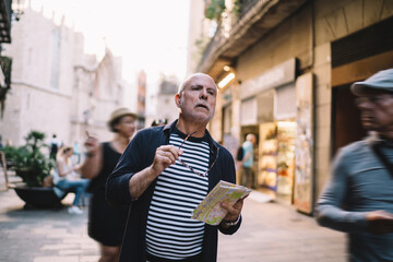 Contemplative male senior with travel map in hand choosing route way for exploring touristic city on vacations, pensive man thoughtful looking at street visiting new town during journey holidays