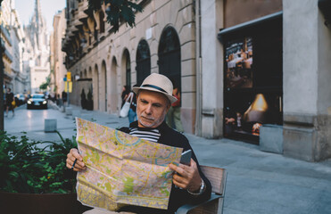 Aged male tourist reading orientation info on location map holding smartphone technology in hand and resting on city bench, Caucasian man in straw hat searching destination during journey holidays