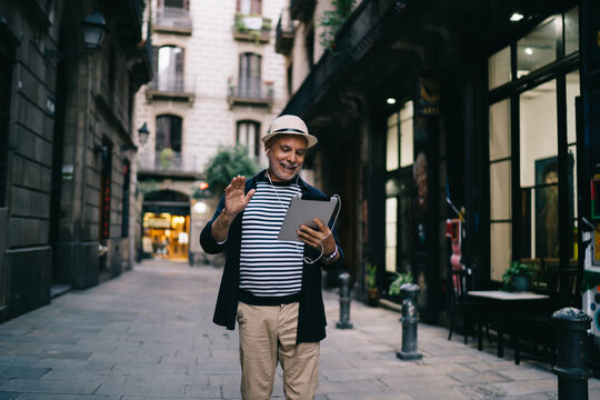 Elderly Caucasian male tourist in electronic headphones making international video call in roaming using modern touch pad and 4g internet, happy man 60s reading network news on gadget website