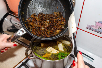 The woman uses a silicone spatula to add fried mushrooms and onion from frying pan to the vegetable soup decoction.