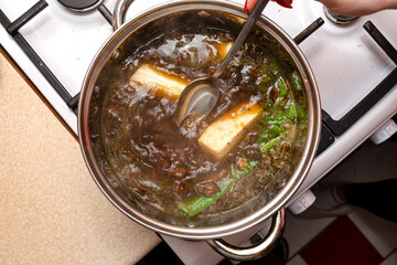 The woman mixes the mushroom soup with a metal spoon which is in a metal pot on the gas stove, top view.