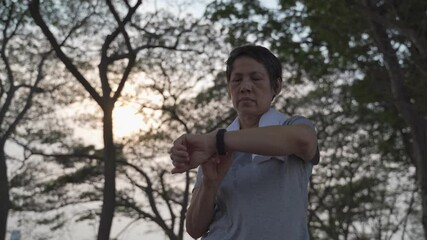 asian senior elderly woman touching smart watch activity tracker at park under tree. finger tapping sport watch for calories and heart rate information. slow motion orbit shot. health care concept. - Powered by Adobe