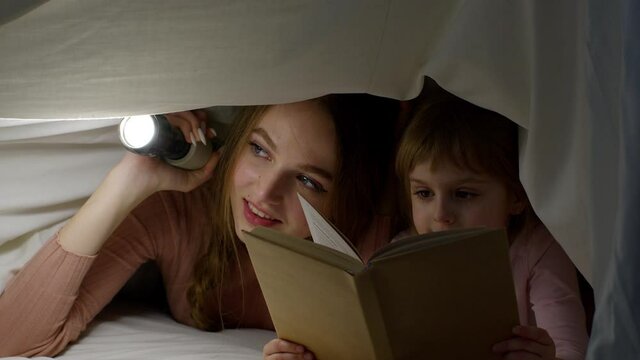 Young mother reading goodnight story fairytale to child daughter under duvet blanket in night room