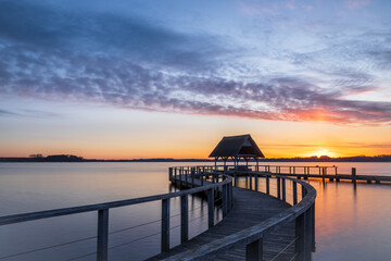 Swinging Pier construction and shelter with thatched roof at beautiful colorful dawn just before sunrise under cloudy sky at Lake Hemmelsdorf, Schleswig-Holstein, Northern Germany