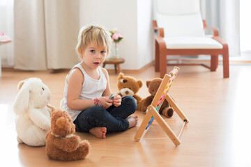 Sweet blond preschool child, toddler boy, playing with abacus at home, surrounded by stuffed toys