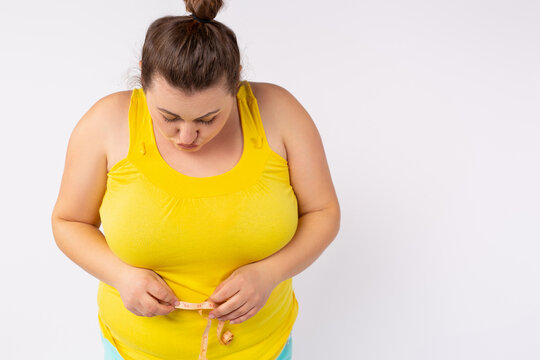 Cropped photo of a plus size woman in a bright T-shirt measuring her belly with a centimeter. Overweight and obesity on a white background.