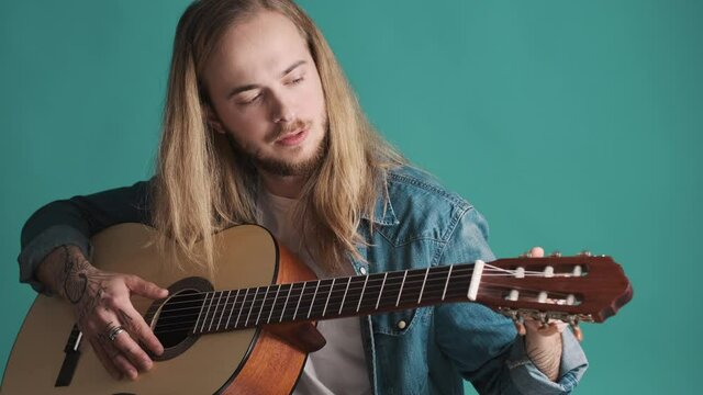 Attractive long haired male musician tuning the guitar isolated on blue background. Young man preparing playing on guitar in studio