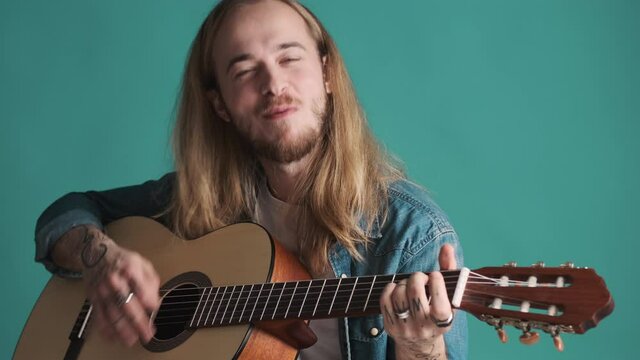 Attractive long haired male musician looking happy playing on guitar and singing isolated on blue background