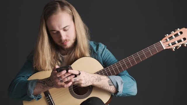Portrait of young bearded male musician with guitar using smartphone during rehearsal isolated on black background. Music concept