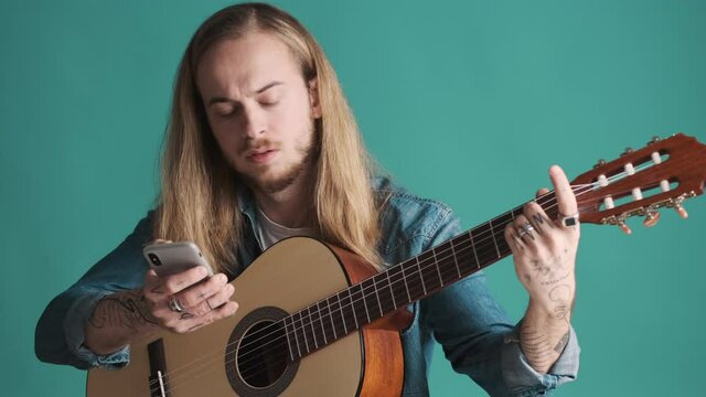 Attractive long haired male musician looking in smartphone how to tuning the guitar over blue background. Music concept