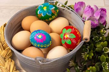 A beautiful Easter, decorative composition with a metal bucket filled with chicken eggs and Easter eggs. Hand-painted colorful Easter eggs. Decorative buckets outdoors with flowers on a wooden backgro