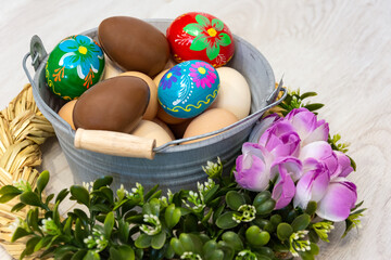 Colorful Easter eggs and chocolate eggs in an Easter basket. Metal bucket full of chicken eggs in a natural color. Bucket surrounded by flowers and a straw wreath.
