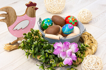 A wooden hen is looking at an Easter colorful decoration with a metal bucket, colorful Easter eggs and a chocolate egg. All surrounded by flowers on a wooden background.