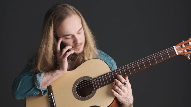Young long haired blond man with guitar talking on smartphone waiting for rehearsal to start over black background