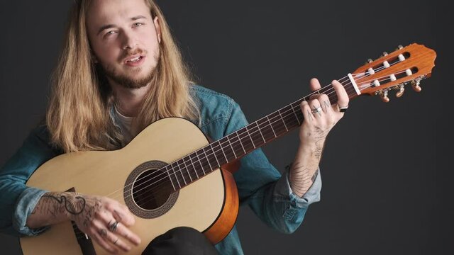 Attractive long haired male musician playing on acoustic guitar and sensually singing on camera isolated on black background