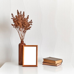 Wooden brown frame mock up, clay vase with brown dried flowers, stack of two old small books on a white coffee table against a white wall background.