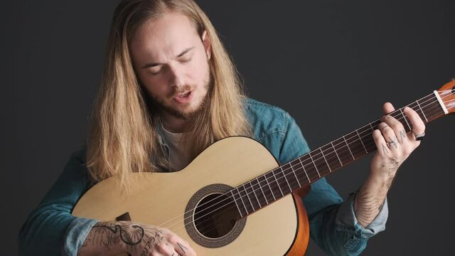 Handsome long haired male musician playing on acoustic guitar singing with eyes closed isolated on black background. Musical instrument concept