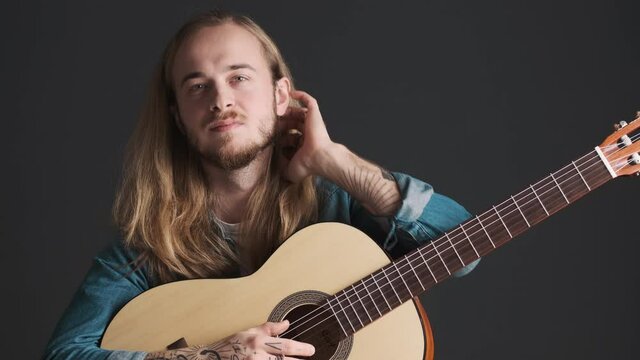 Handsome bearded male musician tucking hair behind ear posing with guitar on camera over black background. Music concept