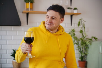 Relaxed young man poses with glass of red wine, standing at kitchen table. Adult guy resting with alcohol in kitchen.